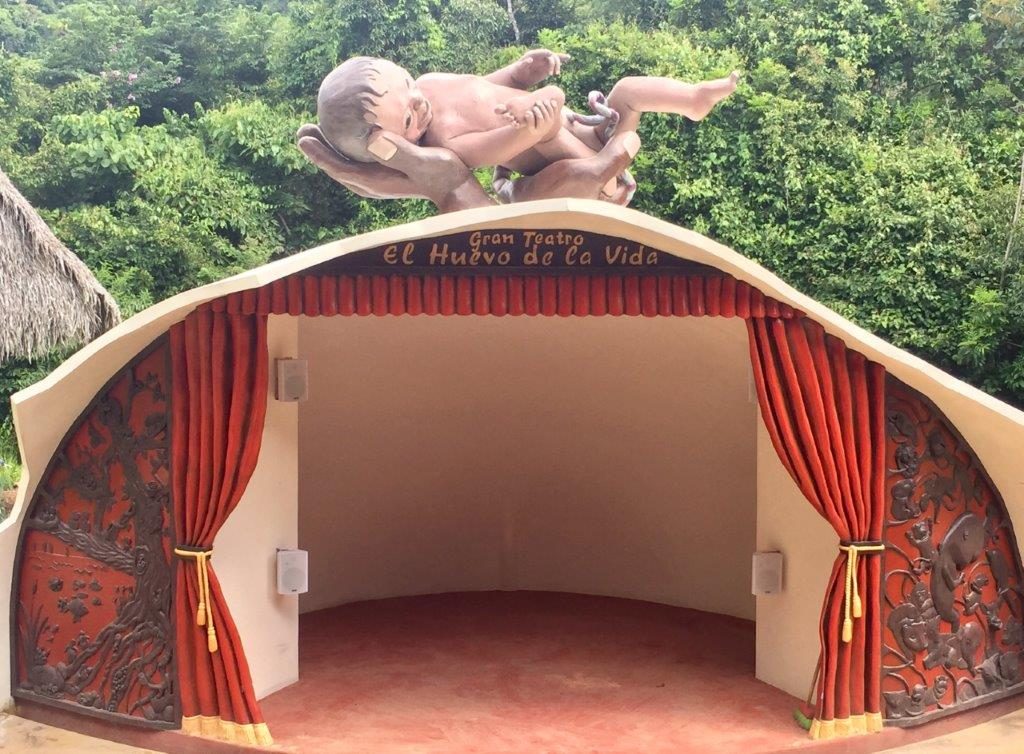 The stage, symbolizing birth, is crowned by two gigantic hands holding a newborn baby, attached to the shell by an umbilical and framed by old theater style red velvet curtains. To the right, there is a wood carving effect of all kinds of newborns, connected by umbilical chords, while on the left another wood carved effect facade depicts scenes of insects, reptiles, birds and other creatures, hatching out of their eggs.